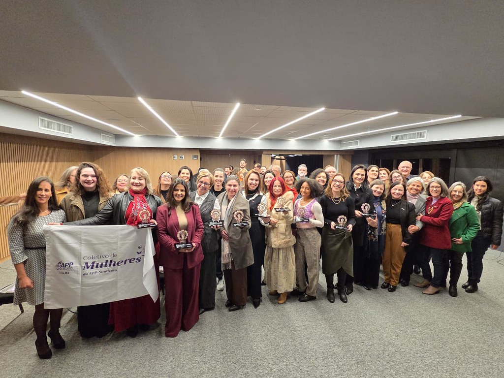 Grupo de mulheres alinhadas em três fileiras dentro de uma sala na sede do Senge-PR. Elas seguram uma bandeira branca do coletivo de mulheres do sindicato.