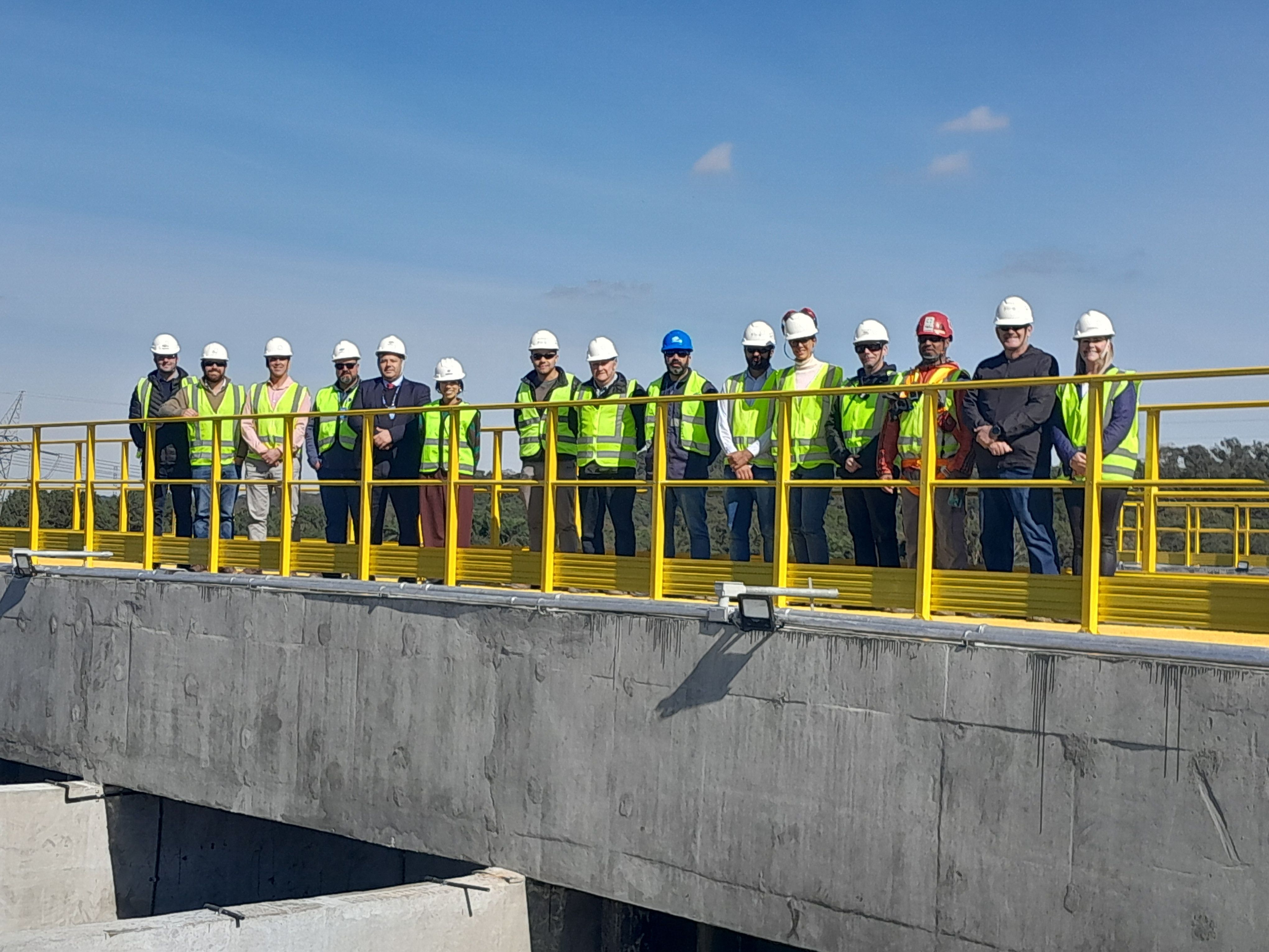 Uma equipe de quinze profissionais e autoridades, vestindo capacetes de segurança e coletes refletivos, posa enfileirada sobre a passarela com guarda-corpo amarelo de uma estrutura de concreto na Estação de Tratamento de Esgoto