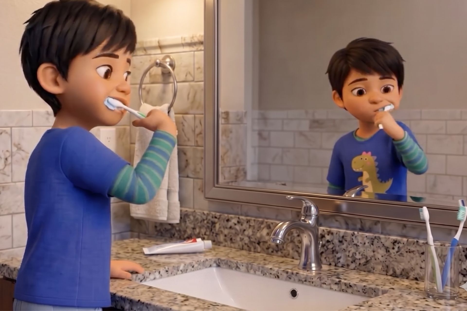 Um menino de cabelo curto e camiseta azul escuro com estampa de dinossauro está escovando os dentes em um banheiro iluminado. Ele está posicionado de perfil à esquerda, olhando para o seu próprio reflexo em um grande espelho à sua frente. A pia de mármore cinza está limpa e a torneira cromada permanece totalmente fechada enquanto ele escova os dentes.