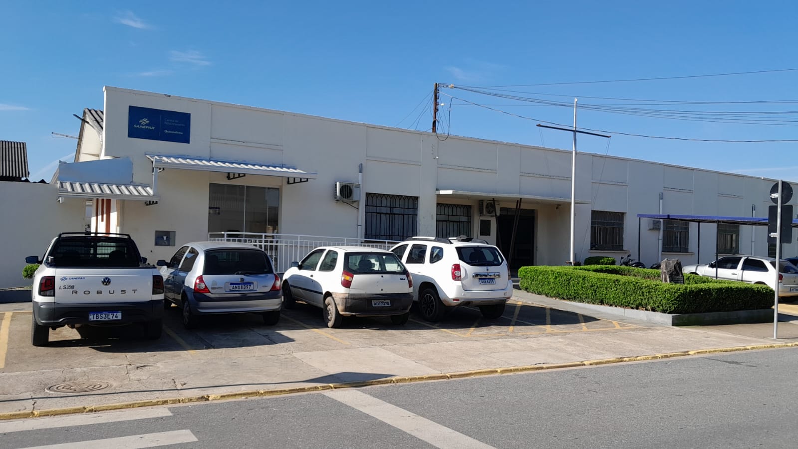 Fachada da Central de Relacionamento da Sanepar em Quitandinha, um prédio térreo branco com placa azul de identificação e veículos estacionados na frente.