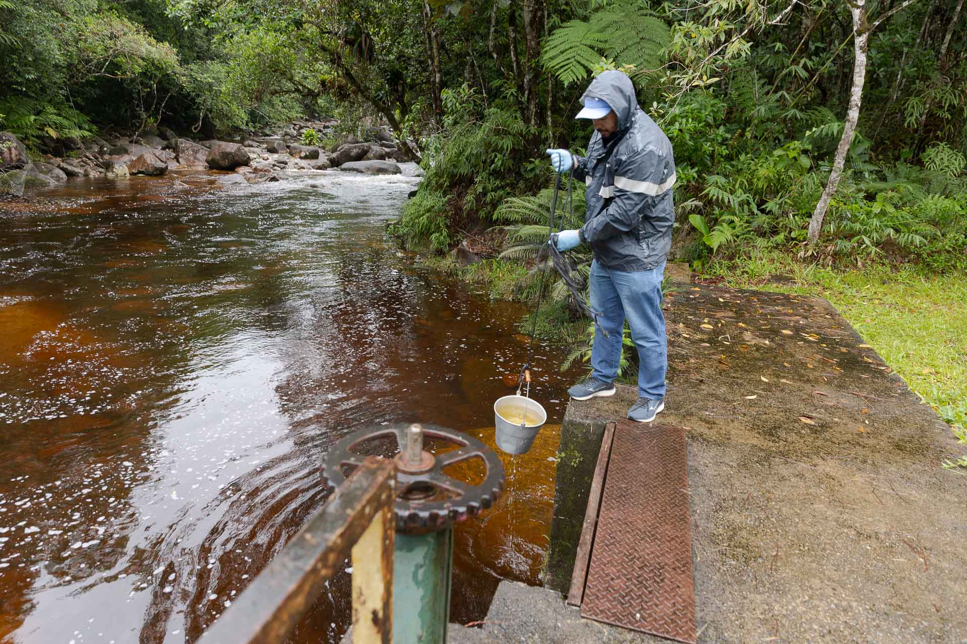 Técnico com capa de chuva e luvas azuis utiliza um balde preso a uma corda para coletar amostra de água em manancial cercado por vegetação.