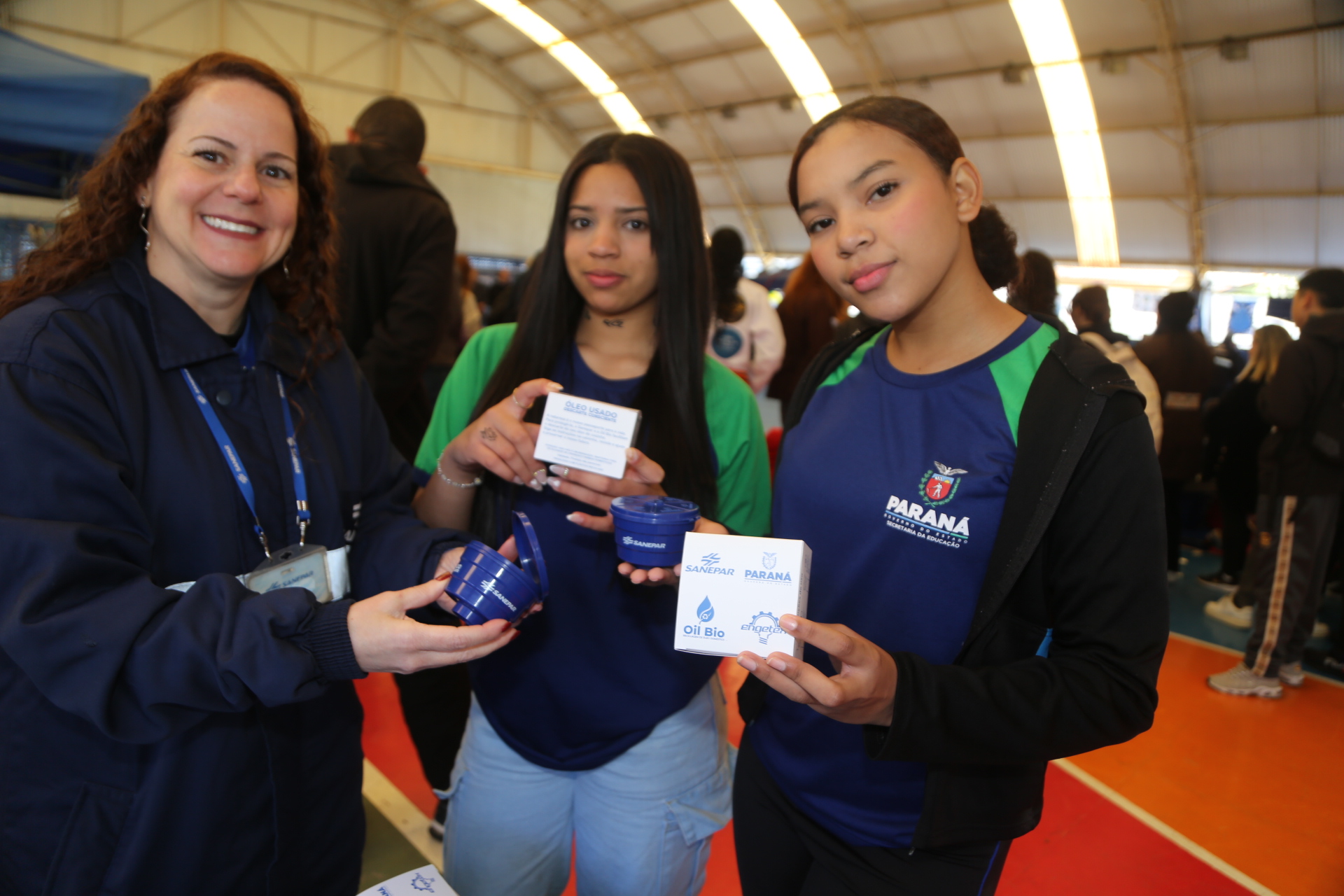 Uma empregada da Sanepar e duas estudantes seguram pequenos coletores de óleo usado em formato de caixa d'´água, redonda e azul, distribuídos pela Sanepar.