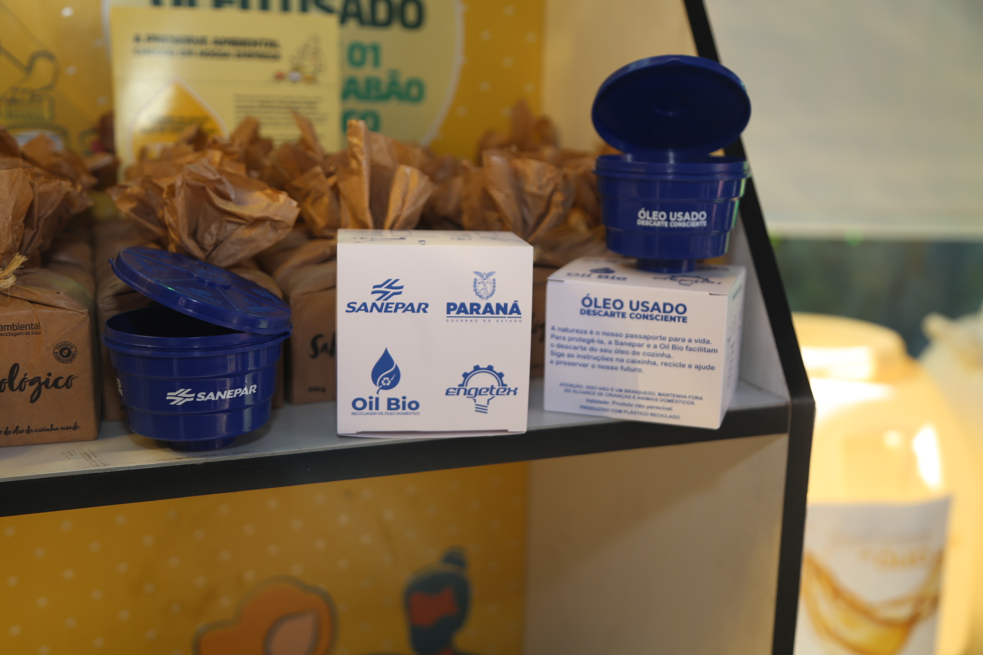 Dois coletores de óleo de cozinha usado em formato de caixa d'água, redondos e da cor azul escura, são exibidos ao lado de suas embalagens, de papelão e brancas. Os itens estão alinhados e possuem a logo da Sanepar