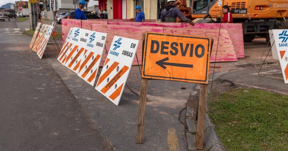 Placa de sinalização amarela com uma seta para a esquerda e o texto "DESVIO". À esquerda, cavaletes zebrados em laranja e branco com a inscrição "OBRAS SANEPAR" delimitam a via interditada. Ao fundo, operários com capacetes e uma escavadeira atuam na obra.