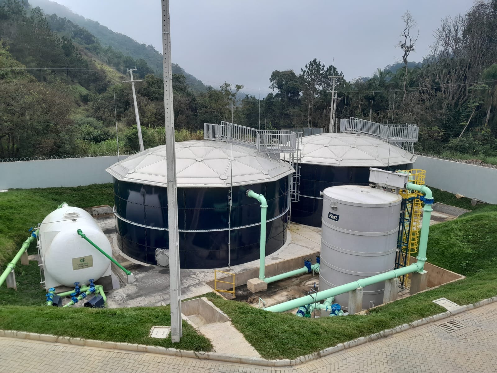 Vista aérea de dois grandes tanques pretos cilíndricos com coberturas metálicas circulares, ladeados por tanques menores e tubulações verdes claros em uma área gramada.