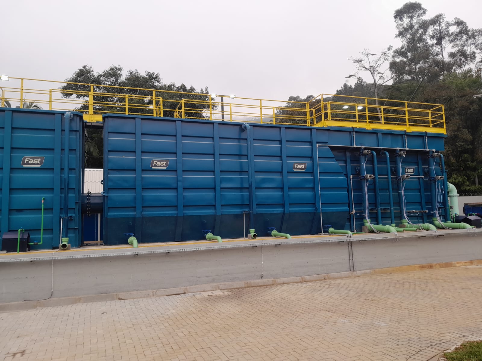 Uma grande estrutura modular metálica pintada de azul-escuro com guarda-corpos amarelos no topo, exibindo tubulações horizontais verdes instaladas na base de concreto.