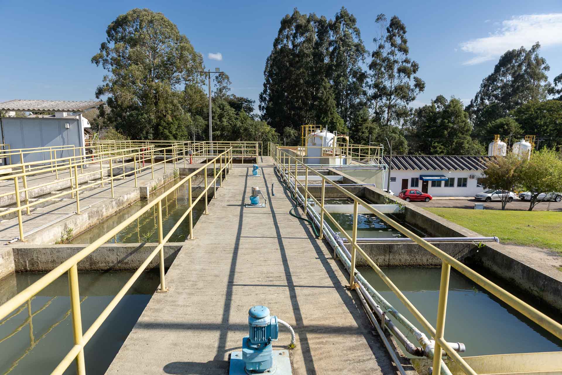 Passarela central da estação de tratamento com motores azuis de bombeamento, ladeada por tanques de água e estruturas operacionais, com árvores e prédios administrativos ao fundo.