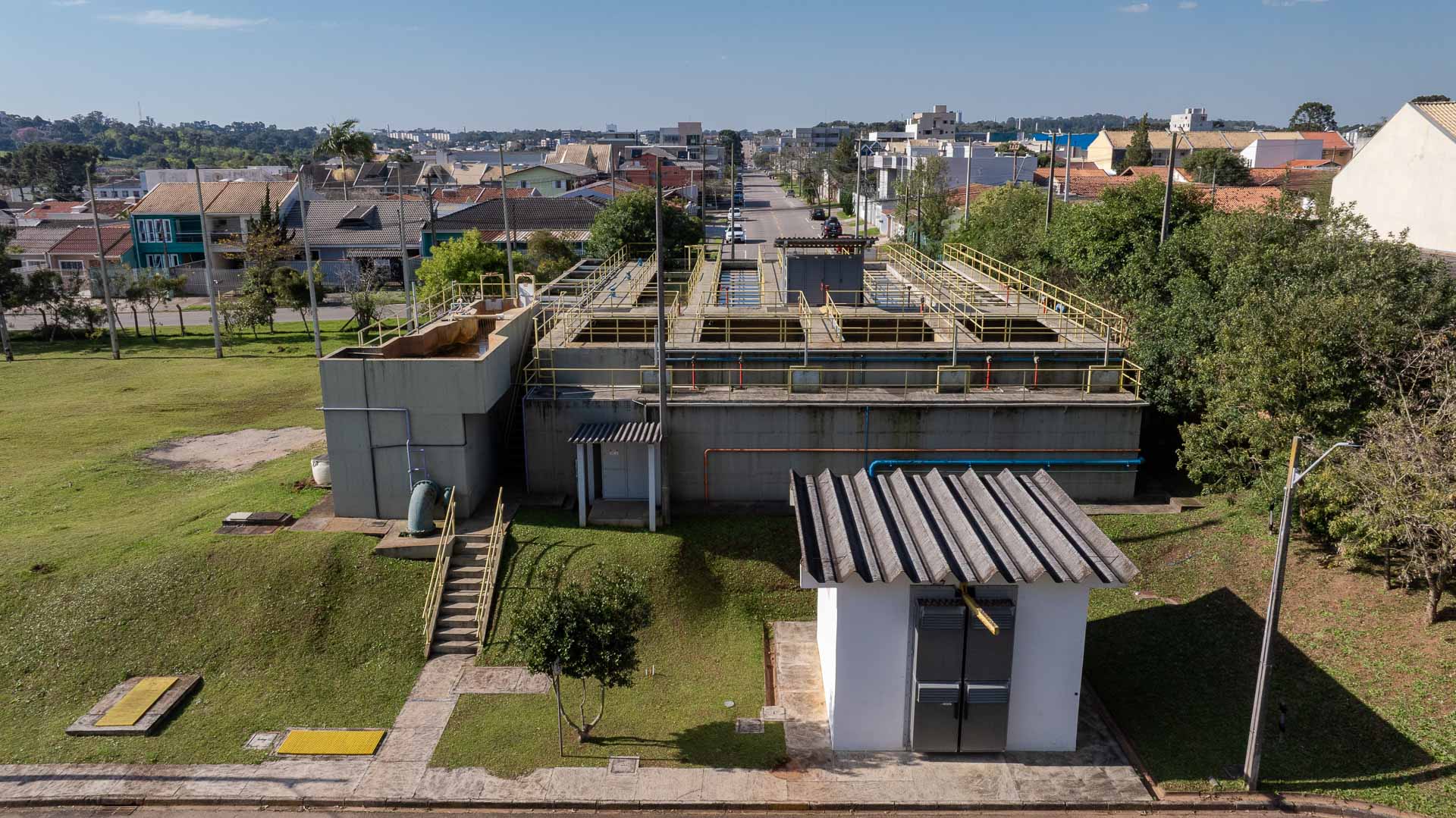 Vista aérea do complexo da Estação de Tratamento de Água (ETA) em Araucária, destacando os tanques de tratamento em concreto e guarda-corpos amarelos, cercados por área verde e casas do bairro ao fundo