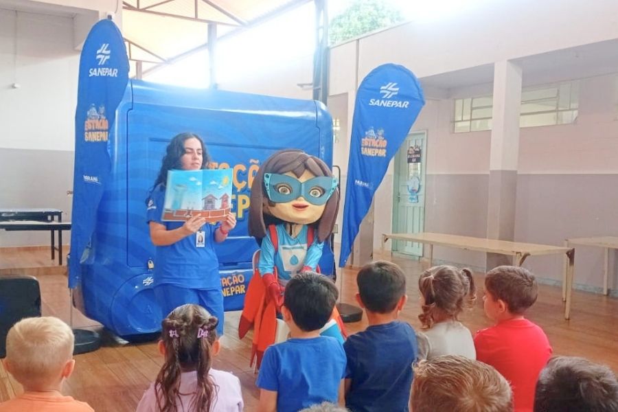 Crianças sentadas no chão assistindo atentamente a uma contação de histórias feita por uma funcionária da Sanepar e pela mascote super-heroína, em frente a uma estrutura azul móvel do projeto Estação Sanepar