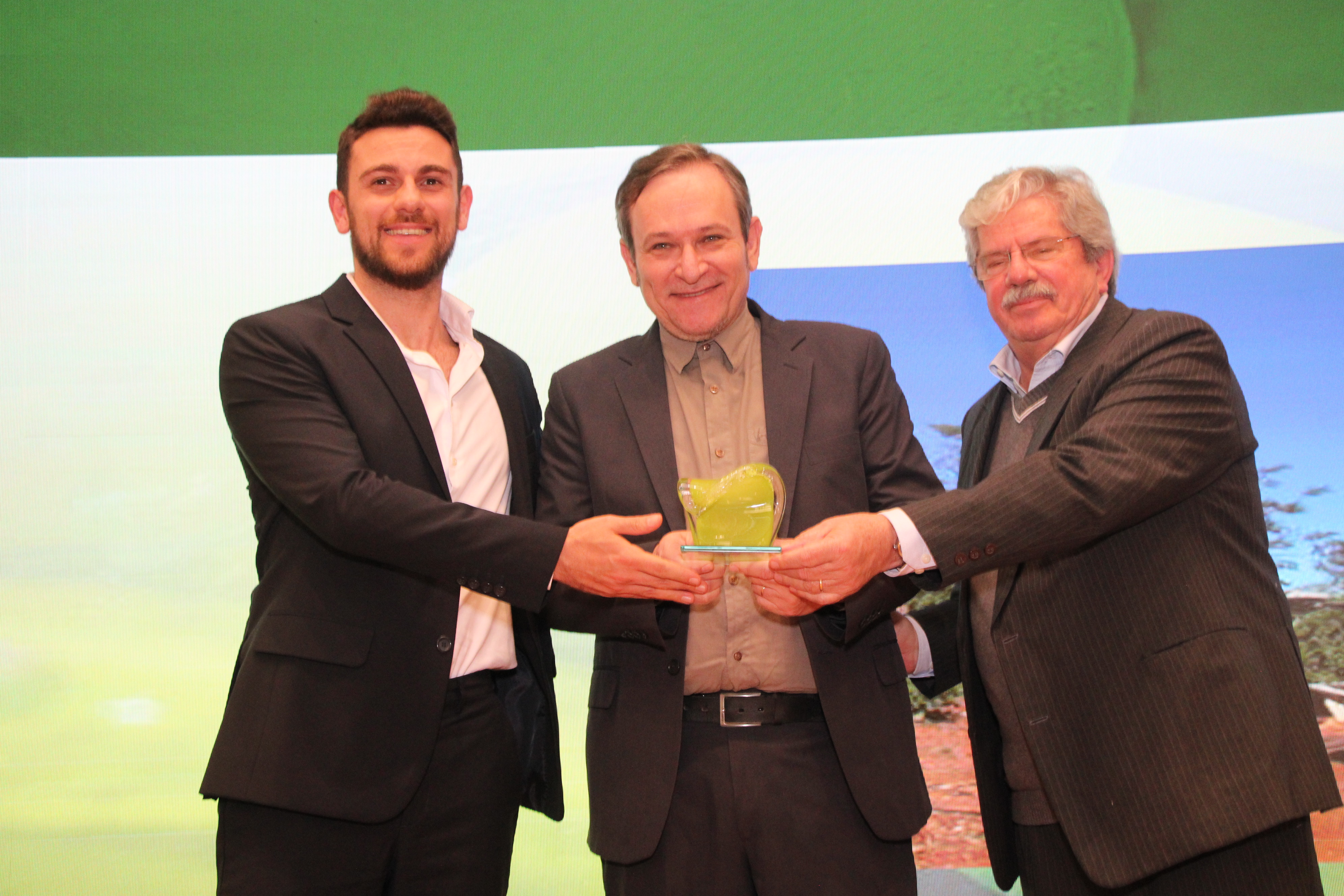 Três homens de terno e camisa social sorriem no palco enquanto o do meio segura um troféu transparente com detalhes verdes.