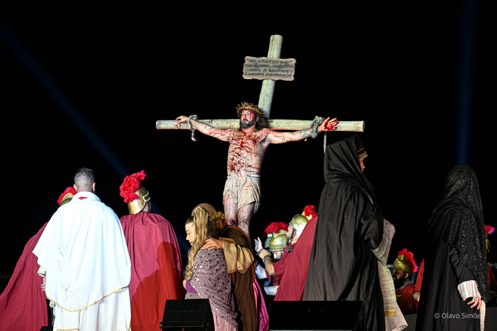 Um ator interpreta a cena do espetáculo A Paixão de Cristo em que Jesus é crucificado. Ele está pregado em uma cruz de madeira e seu corpo está coberto por arranhões com sangue. Ele veste apenas um pano claro que cobre a parte de baixo do seu corpo e usa uma coroa de espinhos. Em cena, ao redor da cruz, também estão atores vestidos como soldados romanos e mulheres vestidas com mantos que cobrem seus cabelos.