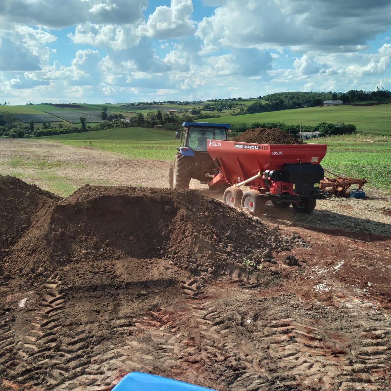 Trator com distribuidor de adubo vermelho sendo abastecido com lodo de esgoto higienizado em uma área agrícola.