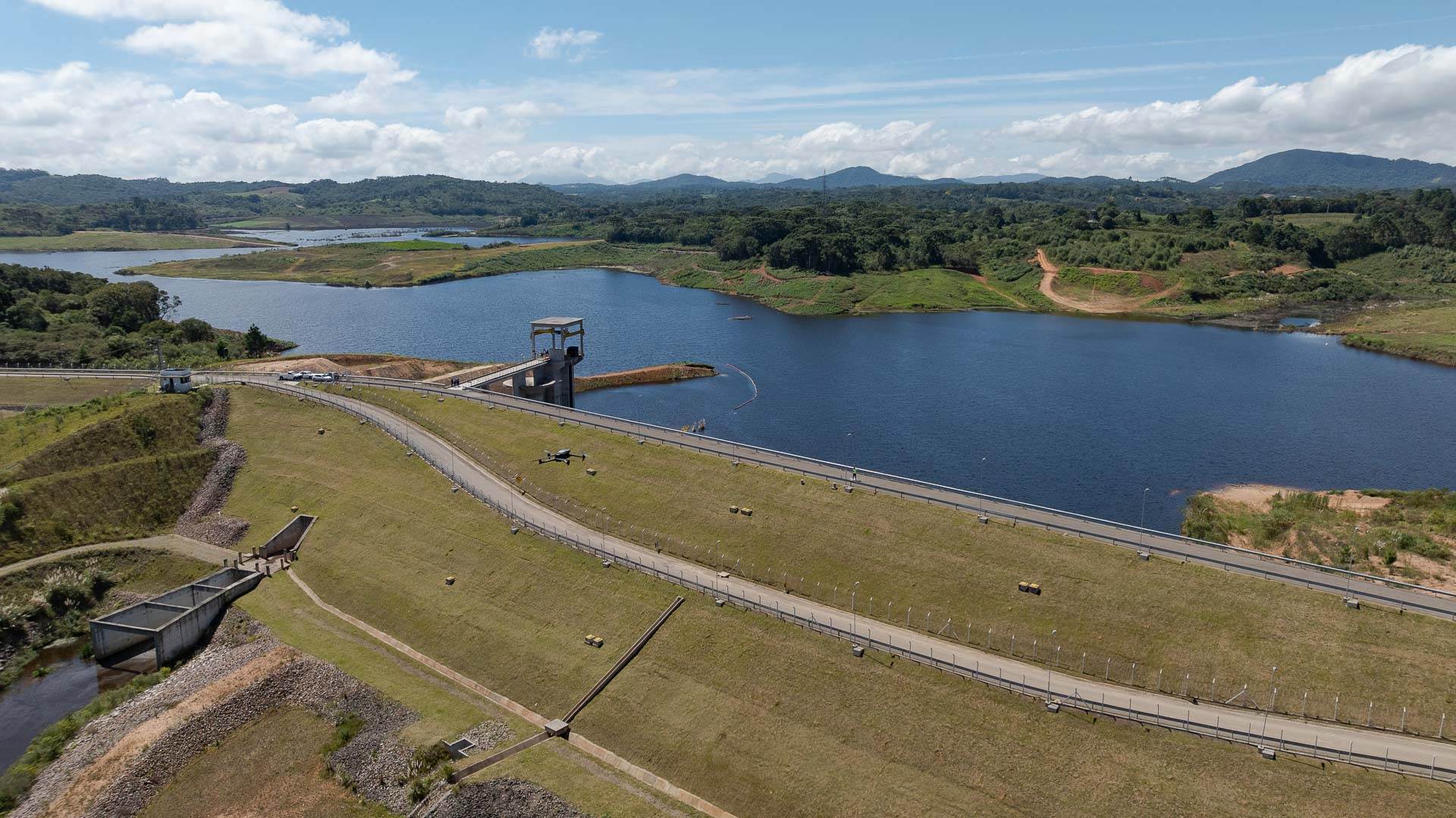 Vista aérea panorâmica da Barragem de Miringuava em um dia ensolarado com nuvens esparsas. À direita, estende-se um grande reservatório de água azul-escura cercado por vegetação nativa, matas e colinas ao fundo. No centro da imagem, o talude de terra da barragem é coberto por grama verde, cortado por vias pavimentadas onde é possível ver pequenos veículos e estruturas técnicas, incluindo uma torre de captação de concreto que avança sobre a água