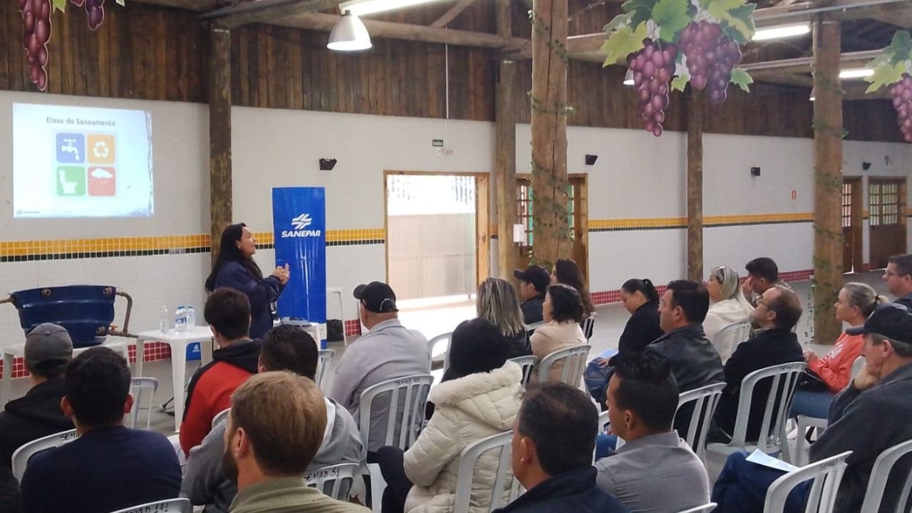 Palestrante da Sanepar realiza palestra sobre eixos do saneamento em espaço comunitário decorado com cachos de uva artesanais, para plateia atenta de moradores.
