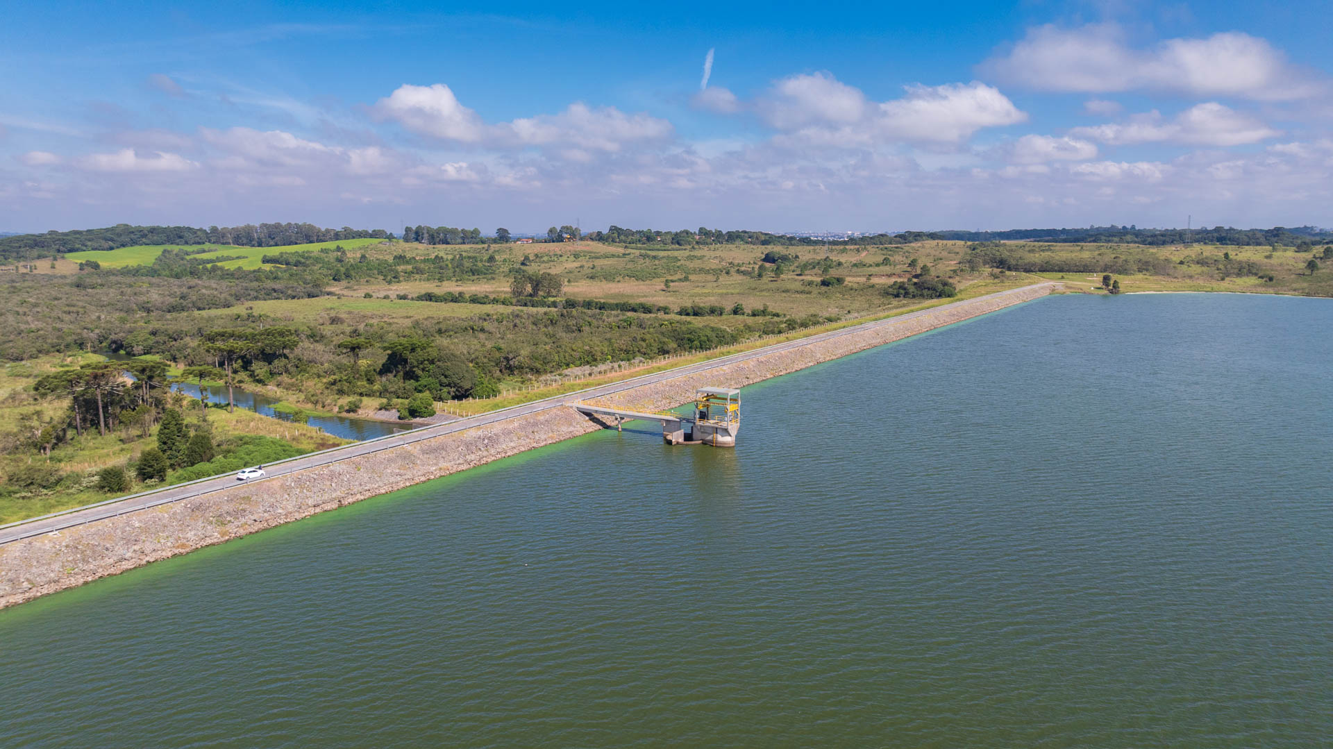 Vista ampla da Barragem do Iraí mostrando o espelho d'água calmo do reservatório, delimitado por uma longa extensão de talude gramado sob o céu com nuvens.