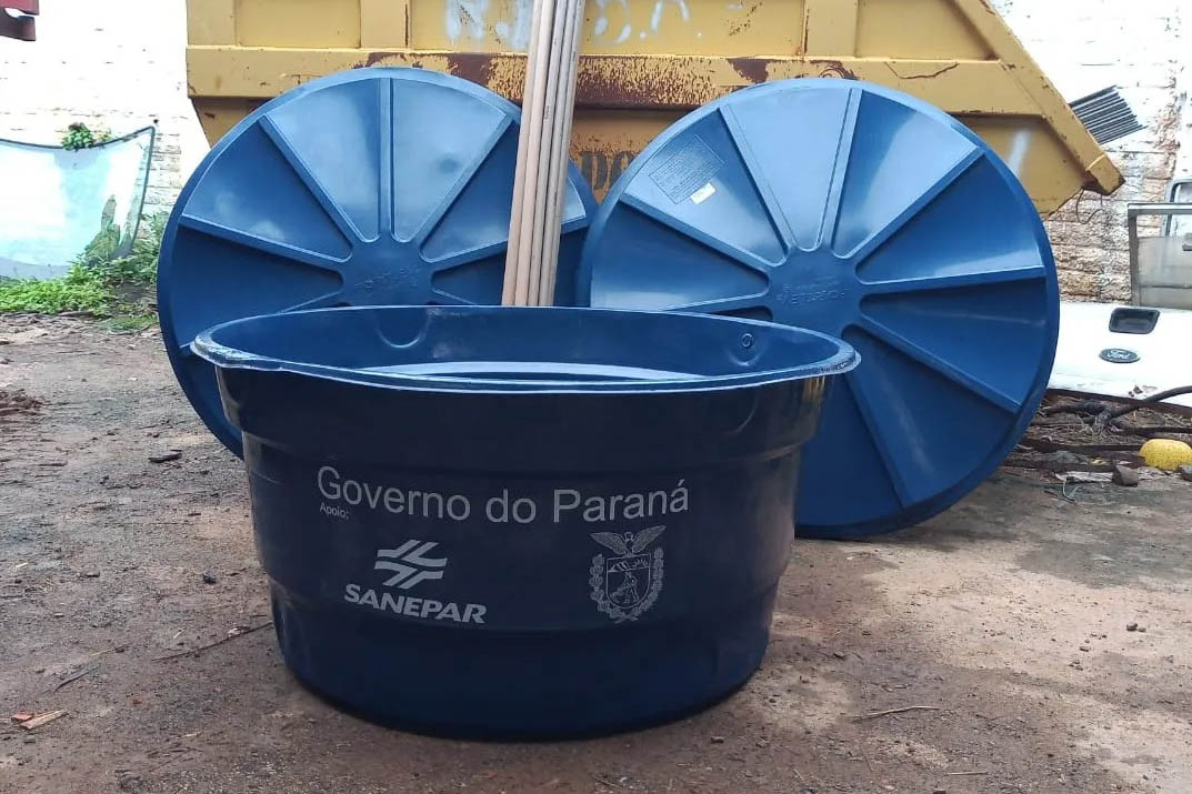 Caixa-d'água azul no chão com as logos do Governo do Paraná e da Sanepar estampadas na frente. Ao fundo, duas tampas azuis estão apoiadas verticalmente contra uma caçamba amarela.