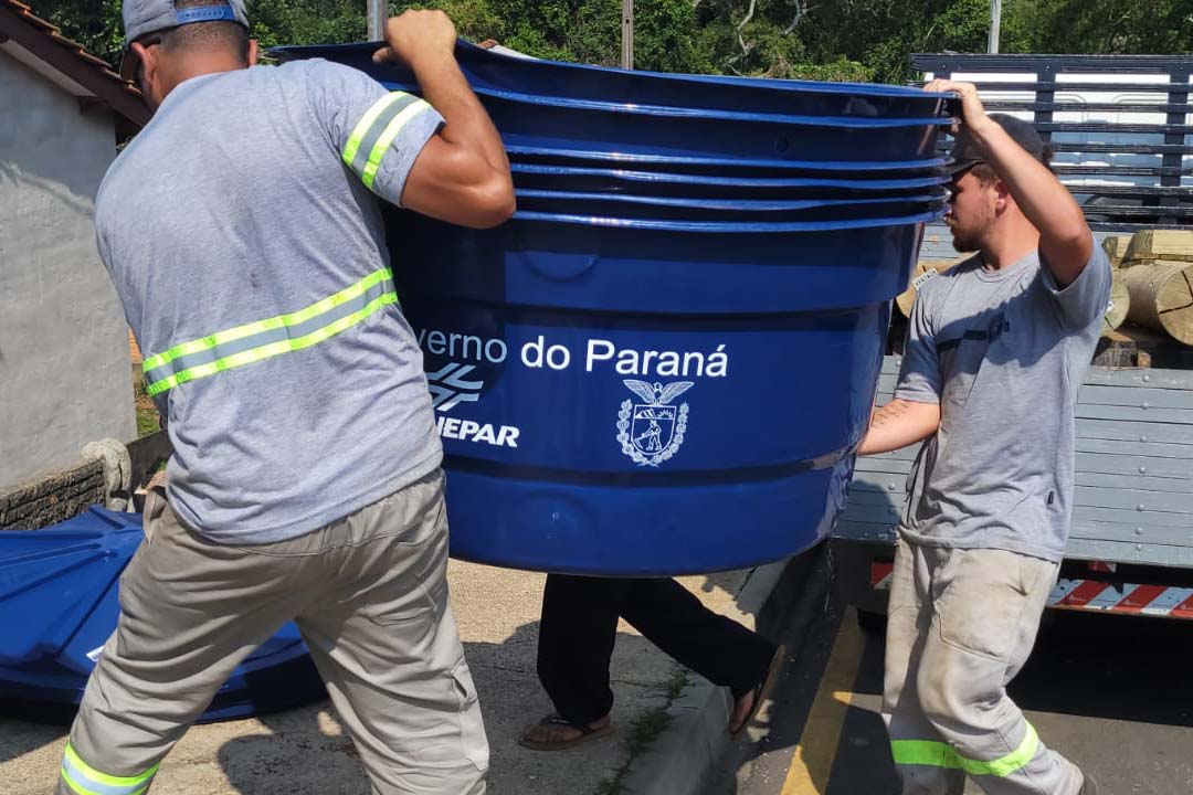 Dois trabalhadores com uniformes cinza carregam uma pilha de caixas d'água azuis. Na lateral da caixa superior, estão estampados o logotipo da Sanepar e o brasão do Governo do Paraná.