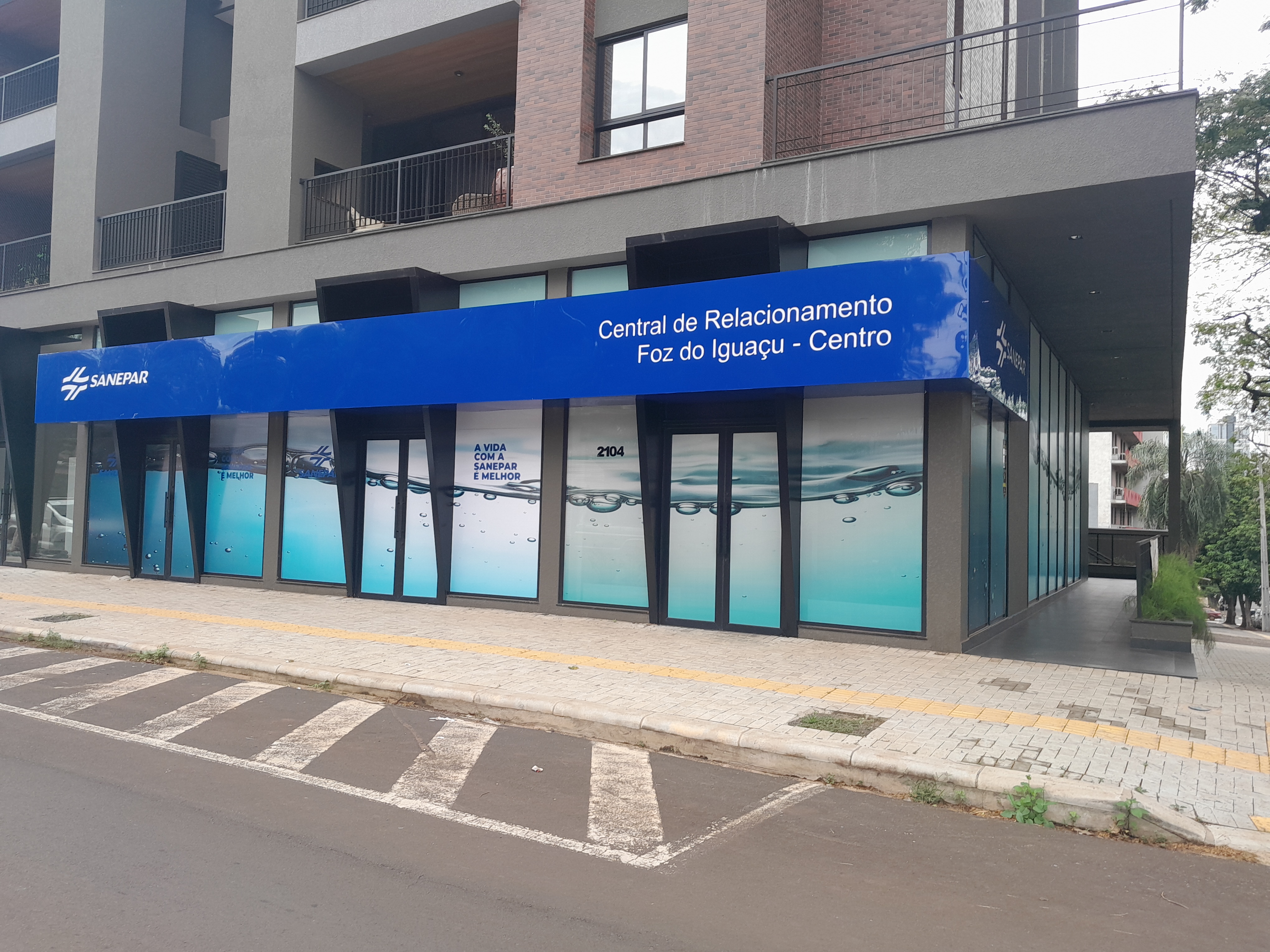 Fachada da Central de Relacionamento da Sanepar em Foz do Iguaçu (Centro), localizada no térreo de um prédio moderno. A fachada possui uma grande faixa azul no topo com a logomarca da Sanepar e os dizeres "Central de Relacionamento Foz do Iguaçu - Centro". As vitrines de vidro exibem o texto "A vida com a Sanepar é melhor" sobre uma ilustração de ondas de água clara