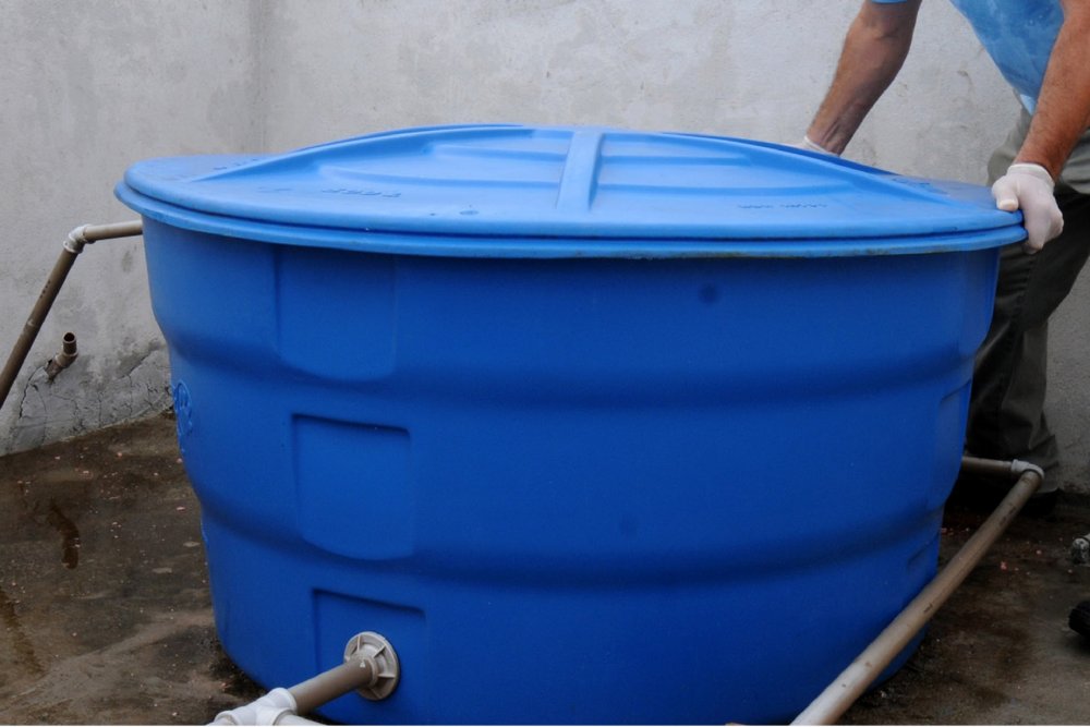 Caixa d'água de plástico azul instalada sobre piso de concreto, com tubulações conectadas à base e uma pessoa de luvas brancas ajustando a tampa.