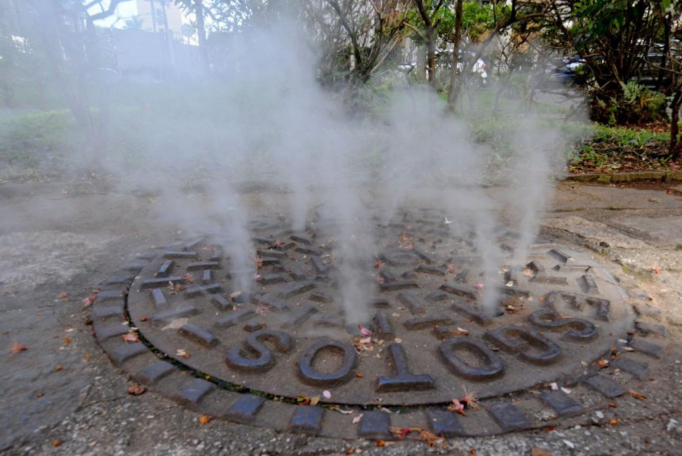 Plumas de fumaça branca saindo através dos orifícios de uma tampa circular de bueiro de esgoto na calçada
