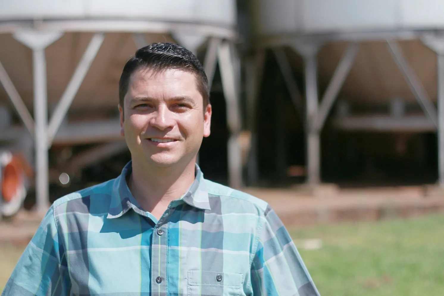 Homem vestindo camisa xadrez á frente de silos de uma área de plantio