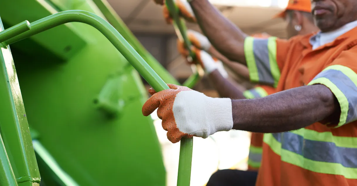 Trabalhadores da limpeza urbana, usando uniforme laranja com faixas refletivas e luvas, seguram o caminhão de coleta de lixo verde durante o serviço.