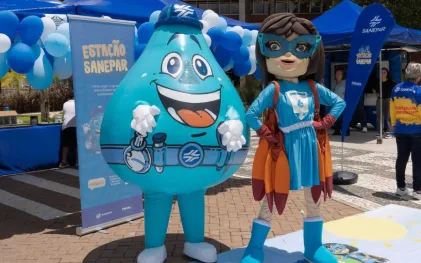 Mascote da Sanepar em formato de gota d'água sorridente e super-heroína com capa e máscara azuis posam lado a lado em evento ao ar livre
