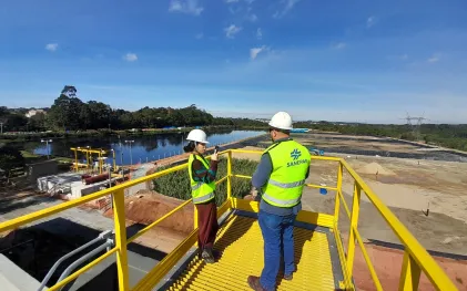 Jornalista entrevista técnico da Sanepar, utilizando capacetes e coletes refletivos, observam a vista panorâmica de uma grande obra e tanques de tratamento a partir de uma plataforma metálica amarela elevada.