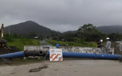 Tubulação azul de grande porte cruza um canal apoiada sobre estrutura de concreto. Operários com capacetes de segurança trabalham na estrutura ao lado de uma retroescavadeira e de uma placa que indica "Em obras Sanepar". Ao fundo, montanhas sob céu nublado.