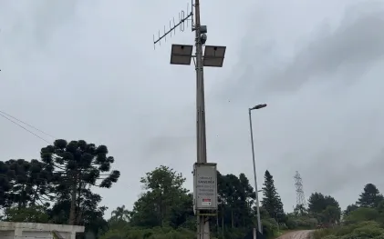 Fotografia em ângulo contrapicado de um poste de concreto que sustenta equipamentos do sistema de alerta da Sanepar, sob um céu nublado e cinzento. Próximo ao topo do poste, há duas placas de energia solar e antenas de transmissão direcionais. Na parte inferior do poste, uma caixa metálica cinza traz inscrições técnicas identificando o equipamento como "REMOTA 01 SANEPAR", acompanhada do telefone de contato 0800 e da indicação "BARRAGEM MIRINGUAVA".