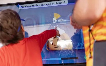Criança toca em tela interativa que exibe a pergunta "Qual é a melhor duração do banho?" com resposta destacada de "5 min"
