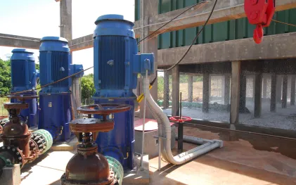 Conjunto de grandes motores elétricos azuis acoplados a tubulações e registros em área operacional. Ao fundo, estrutura de concreto com cortina de água em cascata.