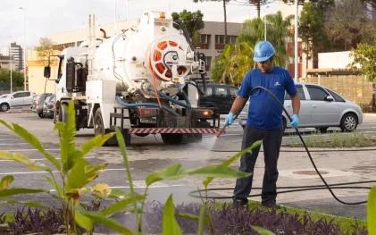 Um empregado da Sanepar (Companhia de Saneamento do Paraná), vestido com uniforme azul, capacete e luvas de proteção, utiliza uma mangueira conectada a um caminhão-tanque branco para regar um canteiro. A operação está sendo realizada com água de reúso.