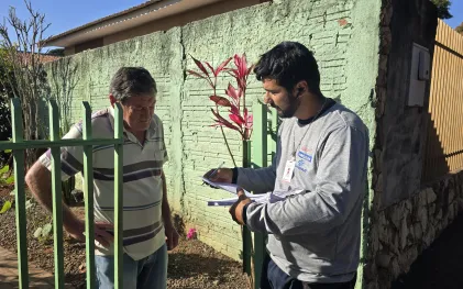 dois homens conversam no portado de uma casa. um é o morador e o outro um agente da sanepar. este está com papeis explicando ao outro como deve proceder para aderir ao serviço de esgoto