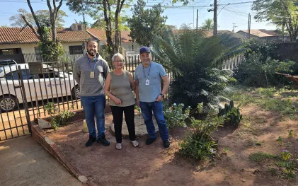 uma senhor de cabelos brancos está no quintal da sua casa, entre dois funcionários da sanepar 