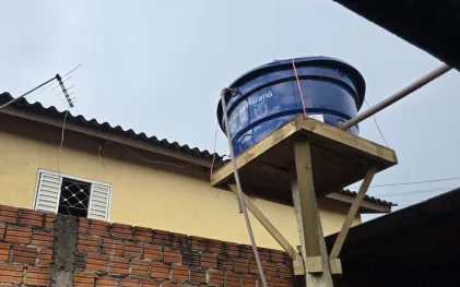 Caixa d'água azul instalada sobre uma estrutura elevada de madeira no quintal de uma residência. No reservatório, destaca-se a inscrição do Governo do Paraná e o logotipo da Sanepar.
