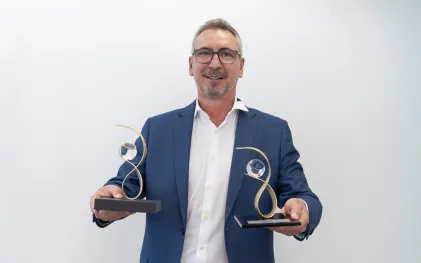 Ozires Kloster está de terno e sorri enquanto segura dois troféus, um em cada mão.