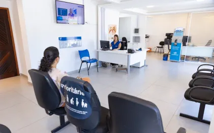 Uma cliente aguarda atendimento em uma Central de Relacionamento da Sanepar. A cliente está sentada em uma das cadeiras enfileiradas em uma recepção. Ao fundo da imagem, é possível ver uma atendente com uniforme azul da Sanepar em frente a um computador. Na imagem também há um painel de senhas exibido em um televisor disposto entre a atendente e a cliente.
