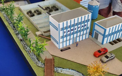 Maquete detalhada que simula uma Estação de Tratamento de Água (ETA) da Sanepar, com prédios administrativos, tanques de decantação, laboratório, reservatório e miniatura de carros.