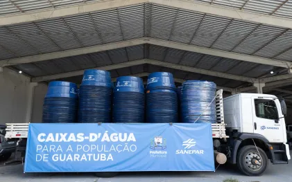 Um caminhão da Sanepar carregado de caixas-d'água azuis empilhadas está estacionado em um galpão. Uma faixa azul na lateral do caminhão tem o texto 'CAIXAS-D'ÁGUA PARA A POPULAÇÃO DE GUARATUBA' e os logotipos da Prefeitura Municipal e da Sanepar.