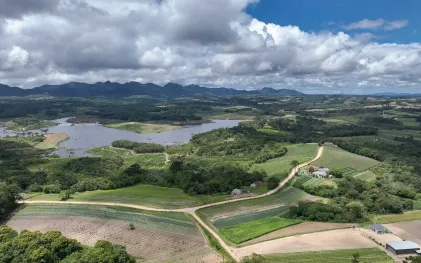 Vista aérea de represa cercada por vegetação nativa e propriedades rurais com áreas agrícolas e estradas de terra, com serras no horizonte sob céu com nuvens.