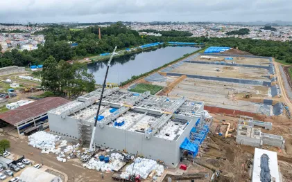 Vista aérea da ampla obra de construção da ETE CIC Xisto, com uma grande estrutura retangular de concreto em destaque, guindaste em operação, lagoa ao fundo e tanques de tratamento biológico por Wetland em andamento