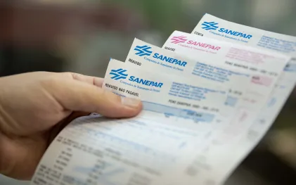 Na foto, uma mão segura quatro faturas impressas com a logo da Sanepar.