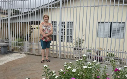 uma senhora loira de cabelos curtos está na frente da sua casa, ao fundo o quintal com flores e a fossa no chão