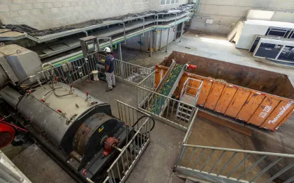 Funcionário opera painel eletrônico de controle em área industrial, ao lado de maquinário mecânico e de uma esteira transportadora que descarta resíduos de alimentos em uma caçamba laranja.