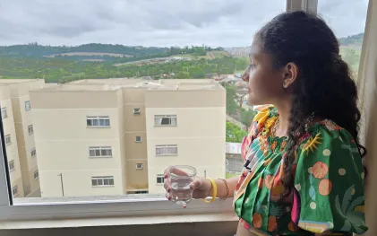 menina sorri com copo de água na mão, na janela de casa