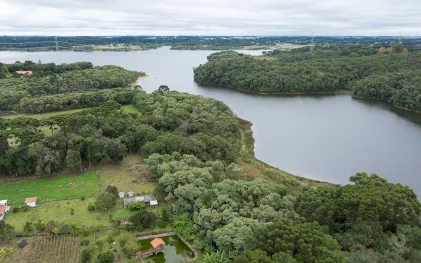 Vista aérea ampla que mostra a sinuosidade de uma represa circundada pela Mata Atlântica. O corpo d'água azul-escuro serpenteia da direita para o centro da imagem, estendendo-se até o horizonte. No canto inferior esquerdo, há uma propriedade rural com um gramado verde-claro bem cuidado, uma casa de dois andares com telhado de cerâmica avermelhada, um pequeno açude retangular com um trapiche de madeira cobertinho e pequenas hortas. A floresta densa, rica em araucárias, abraça toda a margem da represa.