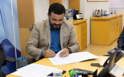 Em primeiro plano, o prefeito de Guaraqueçaba, vestindo paletó claro sobre camisa social azul, aparece sentado à mesa amarela assinando papéis. Ele está sorrindo e concentrado na assinatura.