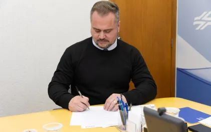 Em primeiro plano, o deputado estadual Alisson Wandscheer, vestindo blusa preta de gola alta sobre camisa clara, aparece sentado à mesa amarela assinando um documento com caneta preta. Ele está concentrado, com o olhar voltado para o papel.