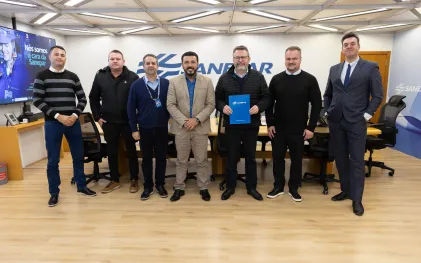 Sete homens alinhados lado a lado em uma sala de reuniões da Sanepar. No centro, o prefeito de Guaraqueçaba, Alessandro Carneiro Soares Truchinski, de paletó claro sorri e, ao seu lado, o diretor-presidente da Sanepar, Wilson Bley, segura uma pasta azul com a marca da Sanepar. Ao fundo, destaca-se o logotipo da companhia na parede.